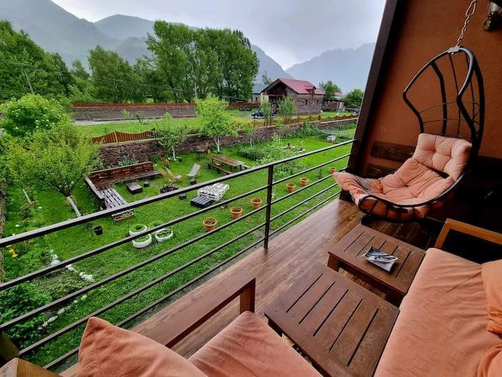 Allegria House Kazbegi — Balcony with hanging chair overlooking garden and mountain scenery.