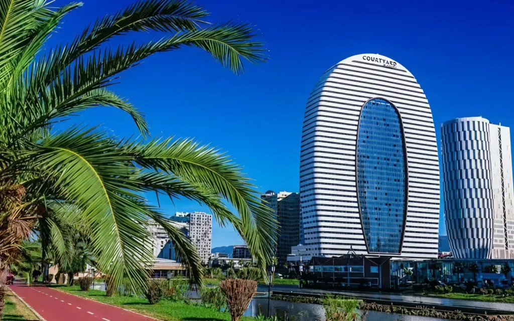 Alliance Palace — Daytime view of the curved high-rise hotel tower beside a palm-lined waterfront promenade.