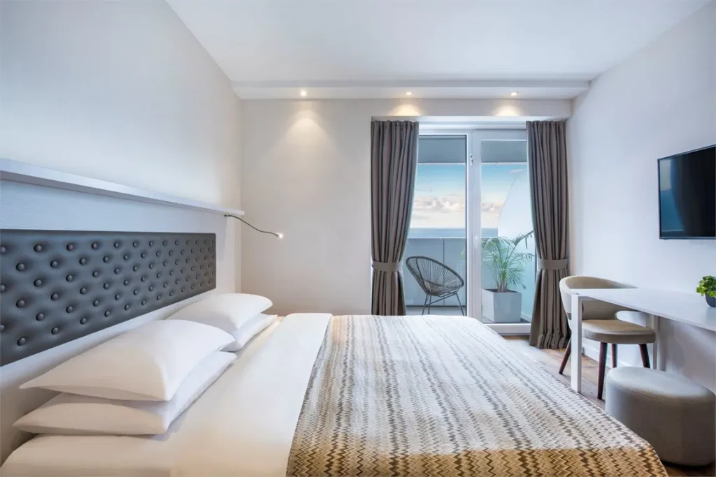 Alliance Palace — Bright hotel bedroom with tufted headboard, balcony access, and a sea view.