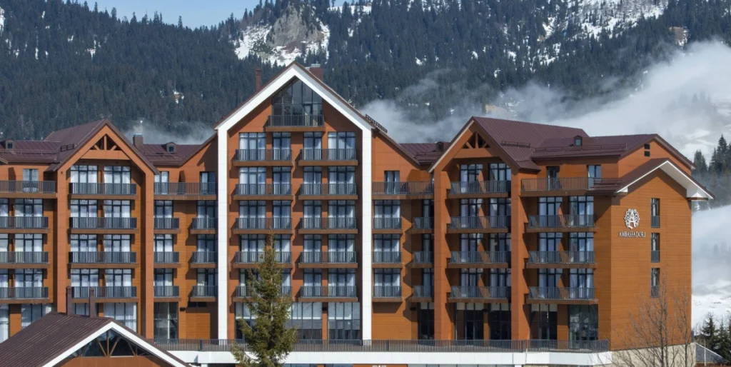 Ambassadori Goderdzi — Hotel exterior with wooden balconies set against snowy mountains and forest.