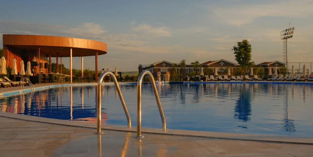Ambassadori Kachreti Golf Resort — Outdoor pool at sunset with poolside loungers, umbrellas, and guest cottages in background.