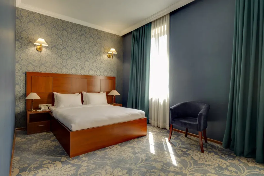 Astoria Tbilisi — Cozy hotel room with wooden headboard, double bed, and teal curtains.