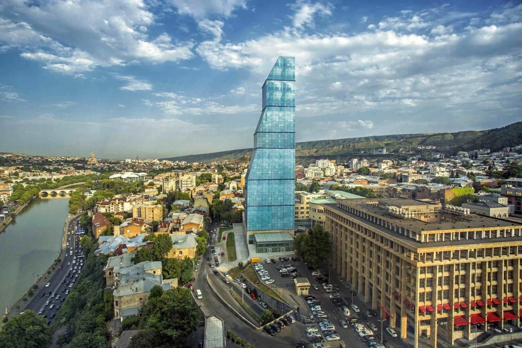 Biltmore Tbilisi Hotel — Aerial view of the hotel's glass tower rising above the Tbilisi cityscape and riverside.