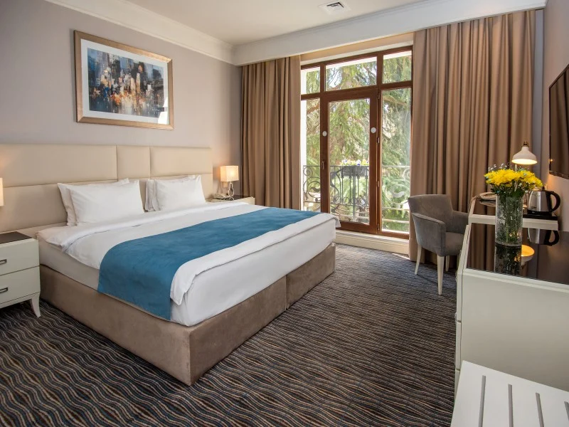 City Avenue Hotel — Bright hotel room with king bed, balcony access, and city-view window.
