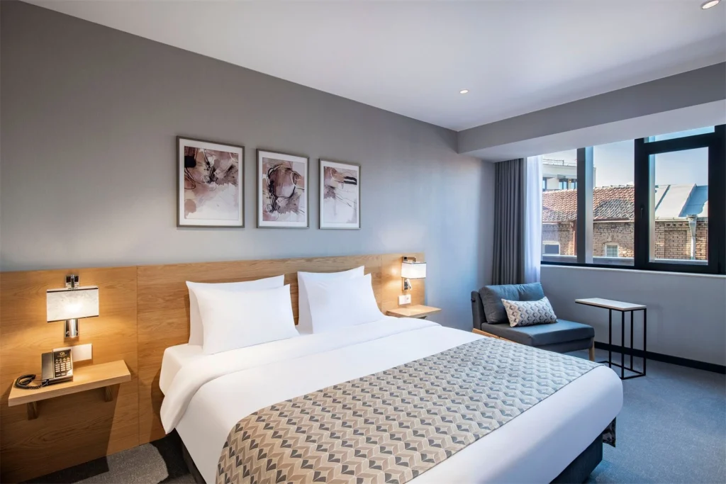Clocks Hotel — Modern guest room with a double bed, armchair, and large window showing rooftops.