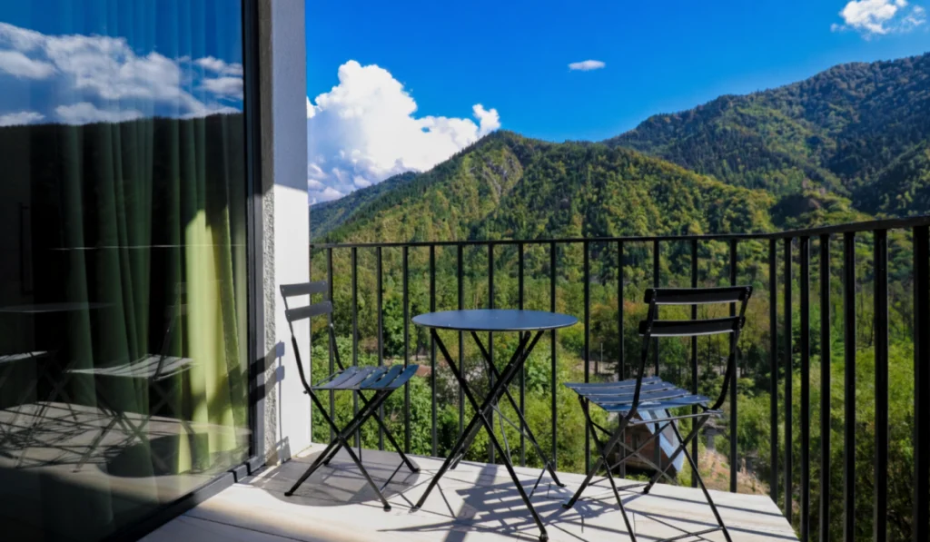Emerald Borjomi — Balcony with table and chairs overlooking green forested mountains under a blue sky.