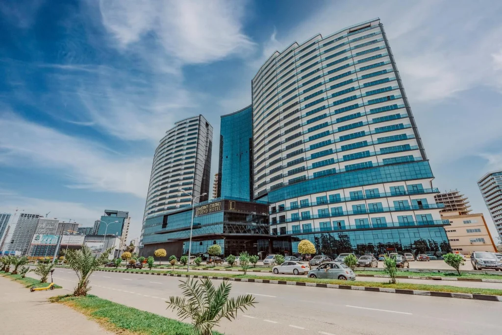 Grand Bellagio Batumi — Tall glass-facade hotel tower with landscaped palm-lined entrance and street view.