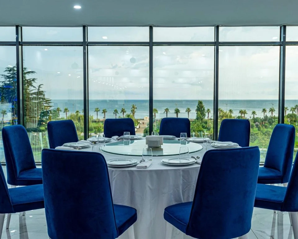 Grand Bellagio Batumi — Round dining table with blue chairs set near floor-to-ceiling windows overlooking the sea.