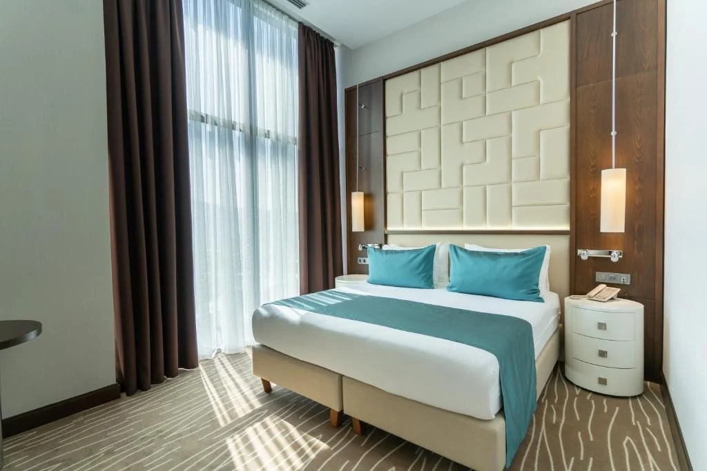 Grand Bellagio Batumi — Hotel room with double bed, teal pillows, and dark wood headboard panel.