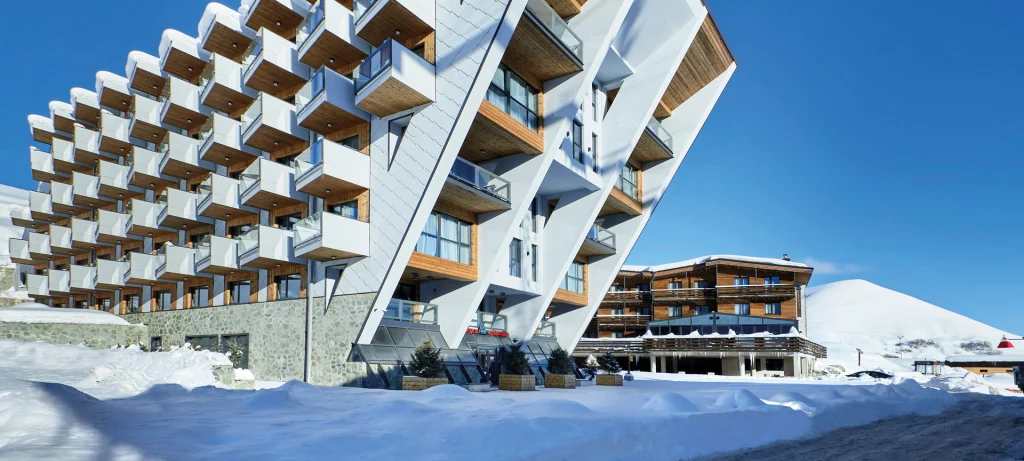 Gudauri Hills Apart Hotel — Modern angular hotel building with balconies surrounded by snow and mountains.