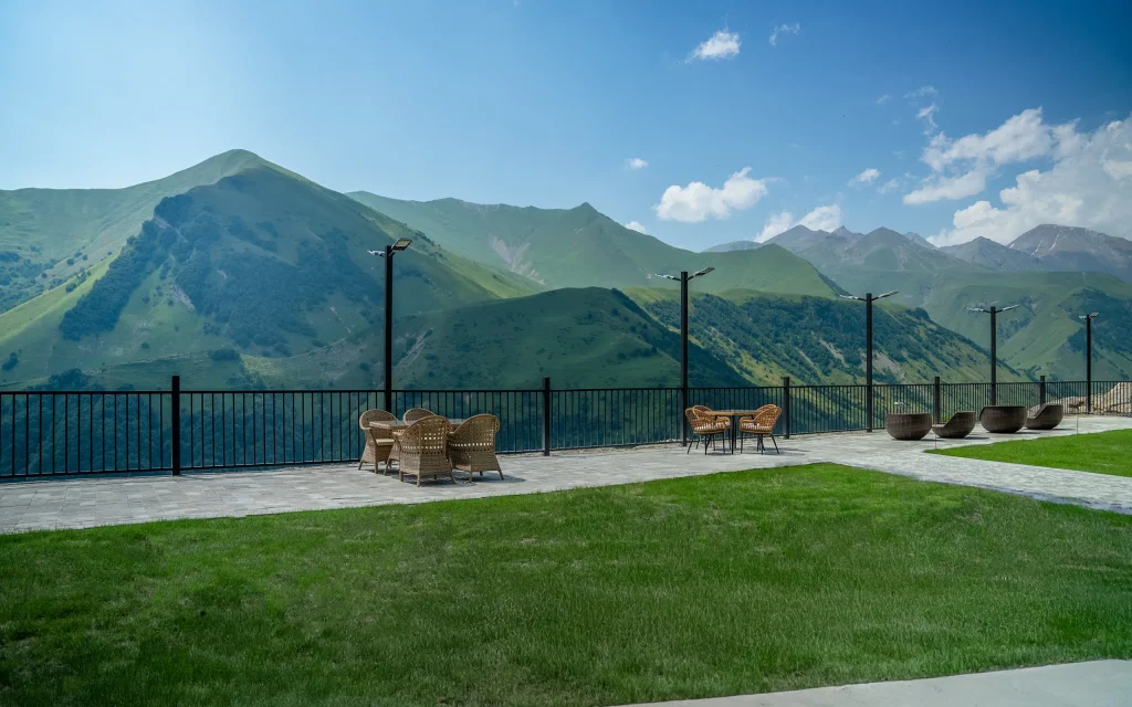 Gudauri Inn — Outdoor terrace with seating and lawn overlooking green mountain ranges under a clear sky.