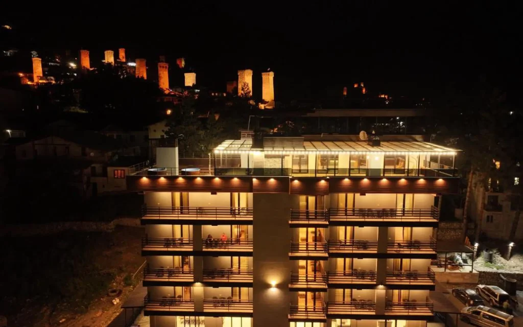 HOTEL USHBA — Night view of the illuminated hotel building with historic towers visible in the background.