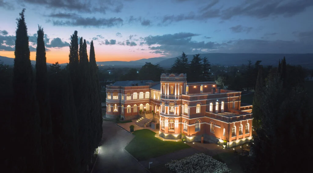 Hameau Mukhrani — Illuminated château-style building at dusk with cypress trees and garden grounds.
