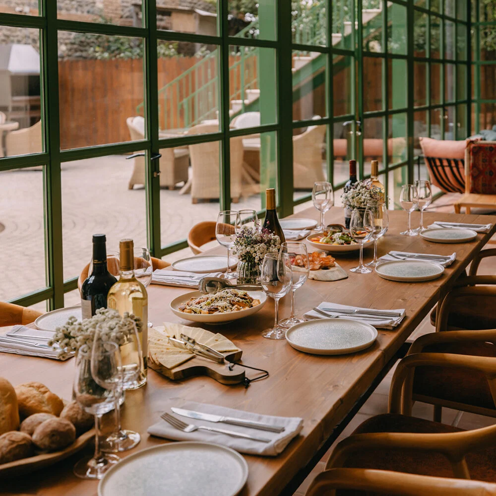 Hameau Mukhrani — Long dining table set with wine and dishes inside a glass-walled restaurant pavilion.
