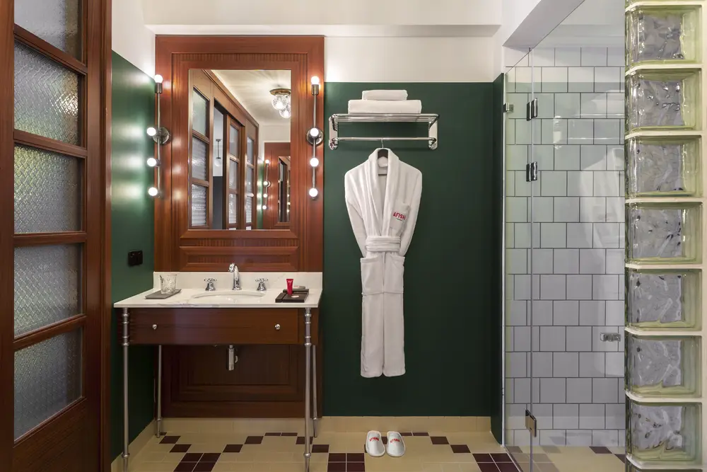 Hotel Afisha — Bathroom with wooden vanity, green walls, glass-block shower, and a hanging robe.