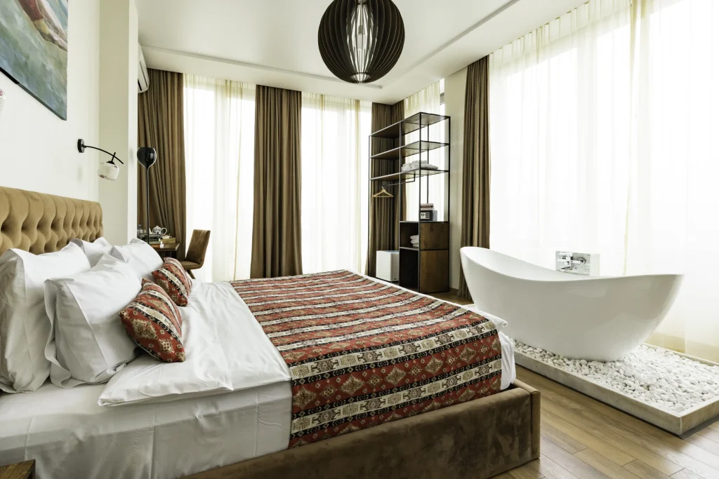 Hotel Flower — Stylish bedroom with patterned bedspread and freestanding bathtub by large windows.