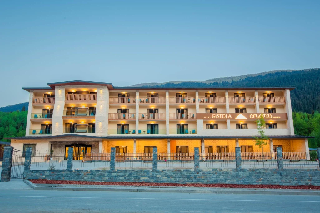 Hotel Gistola In Mestia — Multi-story hotel building with balconies illuminated at dusk against a hillside backdrop.