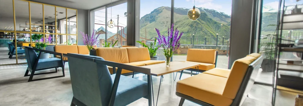 Hotel Memoir Kazbegi — Hotel lounge with yellow and blue armchairs facing large windows overlooking mountains.