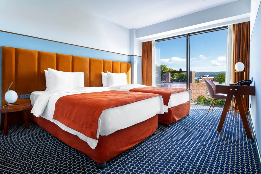 Hotel Zemeli — Twin hotel room with orange headboards, blue carpet and a city view window.
