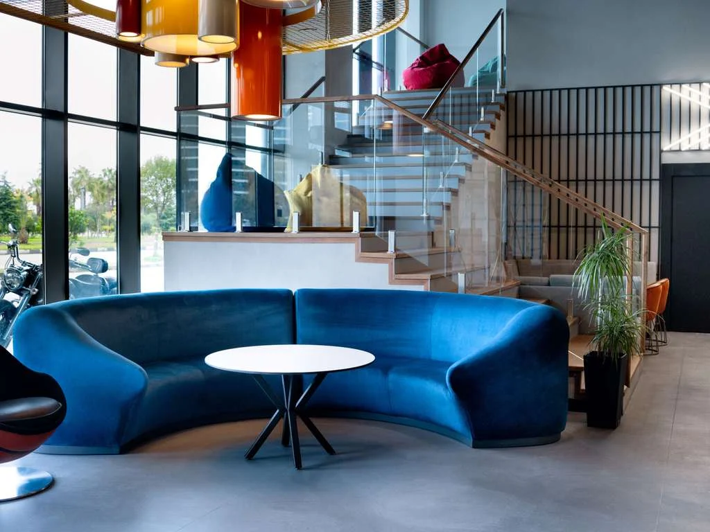 Ibis Styles Batumi — Bright hotel lobby with blue curved sofa, staircase, and floor-to-ceiling windows.