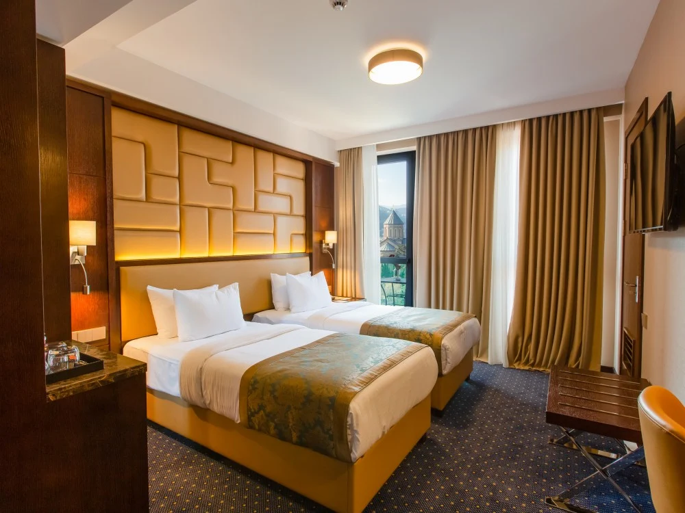 KMM Hotel — Twin beds with upholstered headboard, large window, and city view in the background.