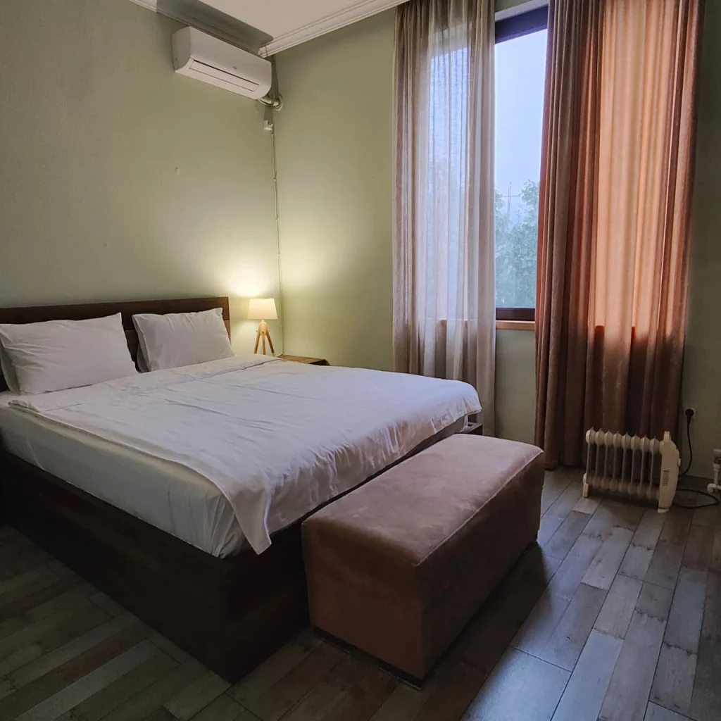 Khvamli 2002 — Bedroom with a double bed, nightstand lamp, and curtained window with natural light.