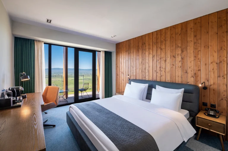 Kvareli Lake Resort — Hotel room with wood paneling, a large bed, and a balcony overlooking the valley.