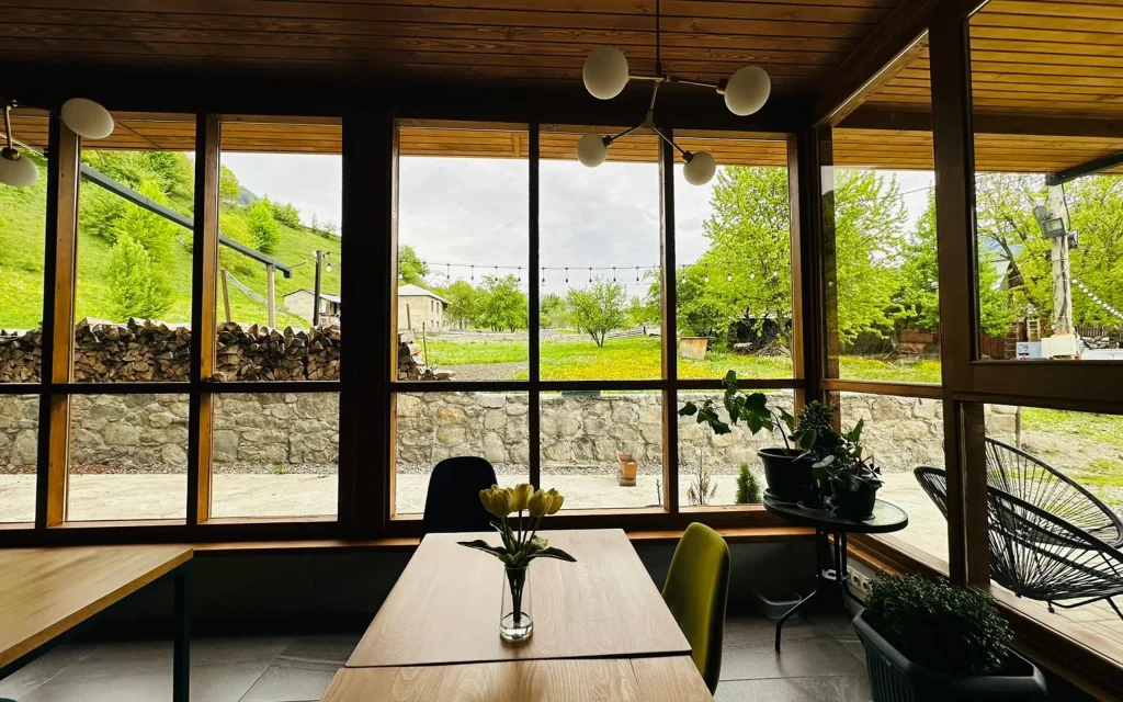 Latour mestia — Glass-walled dining area with wooden tables overlooking a green mountain landscape.