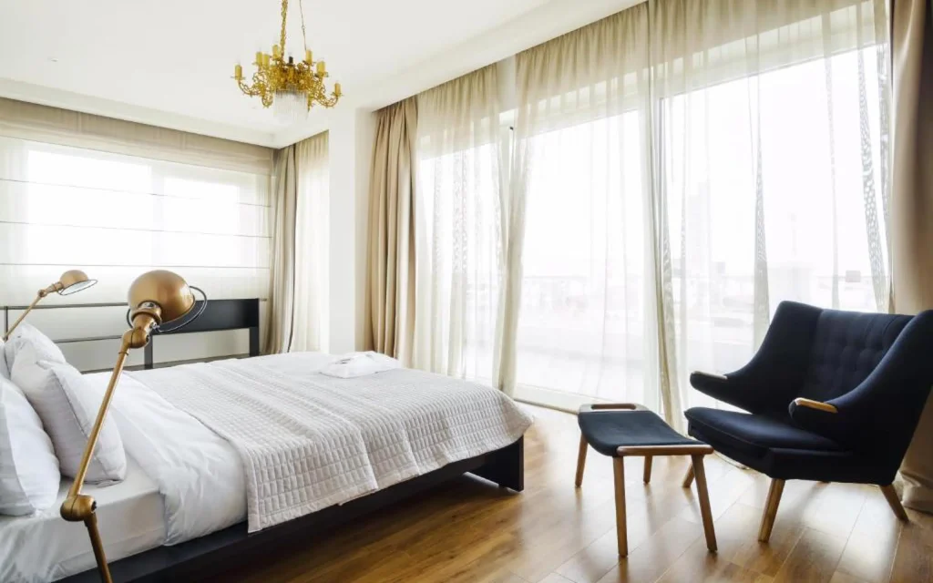 Le Port Boutique Apart Hotel — Bright bedroom with gold chandelier, upholstered chair and large curtained windows.