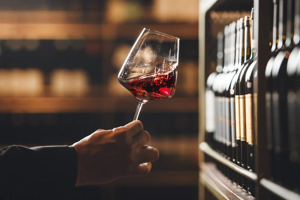 MIDAMO | Wine Village — A hand swirls a glass of red wine near a bottle rack.