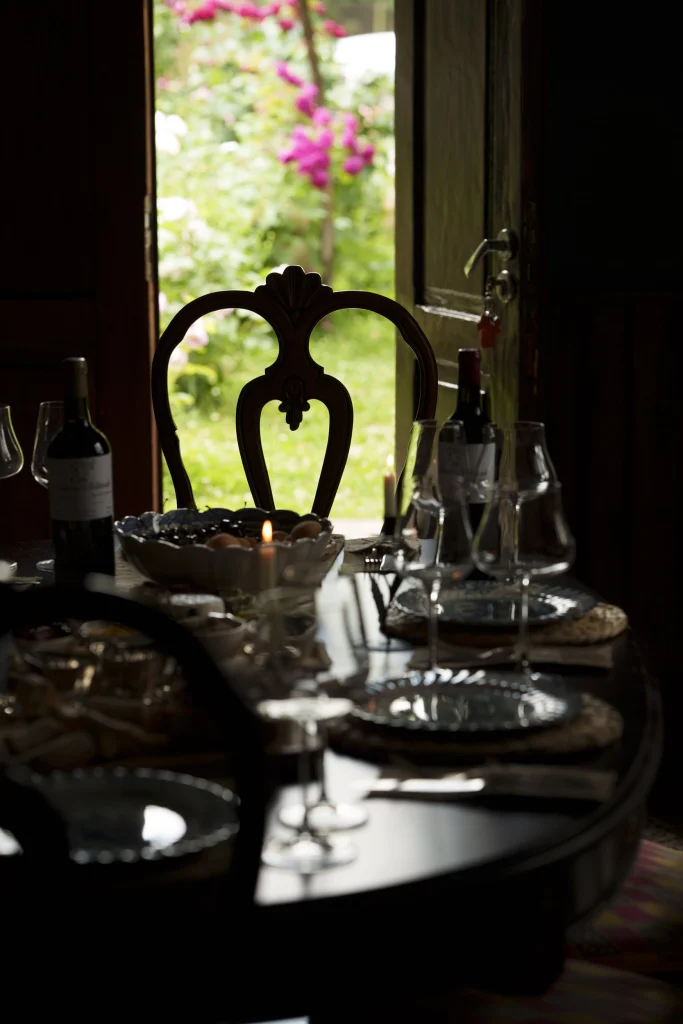 Marani Milorauli — Dim interior dining table with wine bottles and glasses near an open garden door.