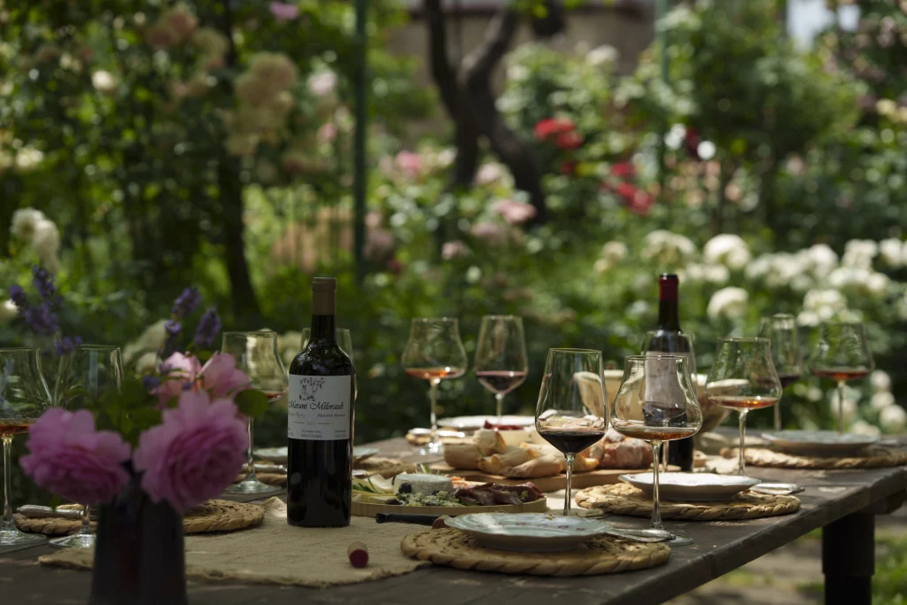 Marani Milorauli — Outdoor dining table set with wine and food amid a lush garden setting.