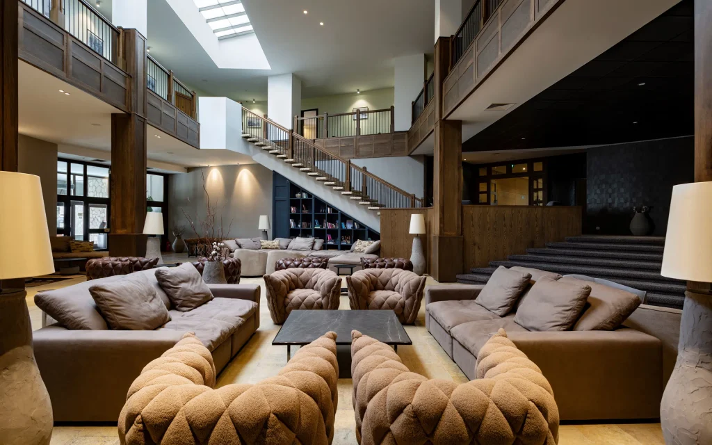 Marco Polo Hotel Gudauri — Spacious hotel lobby with sofas, armchairs, a grand staircase, and wooden accents.