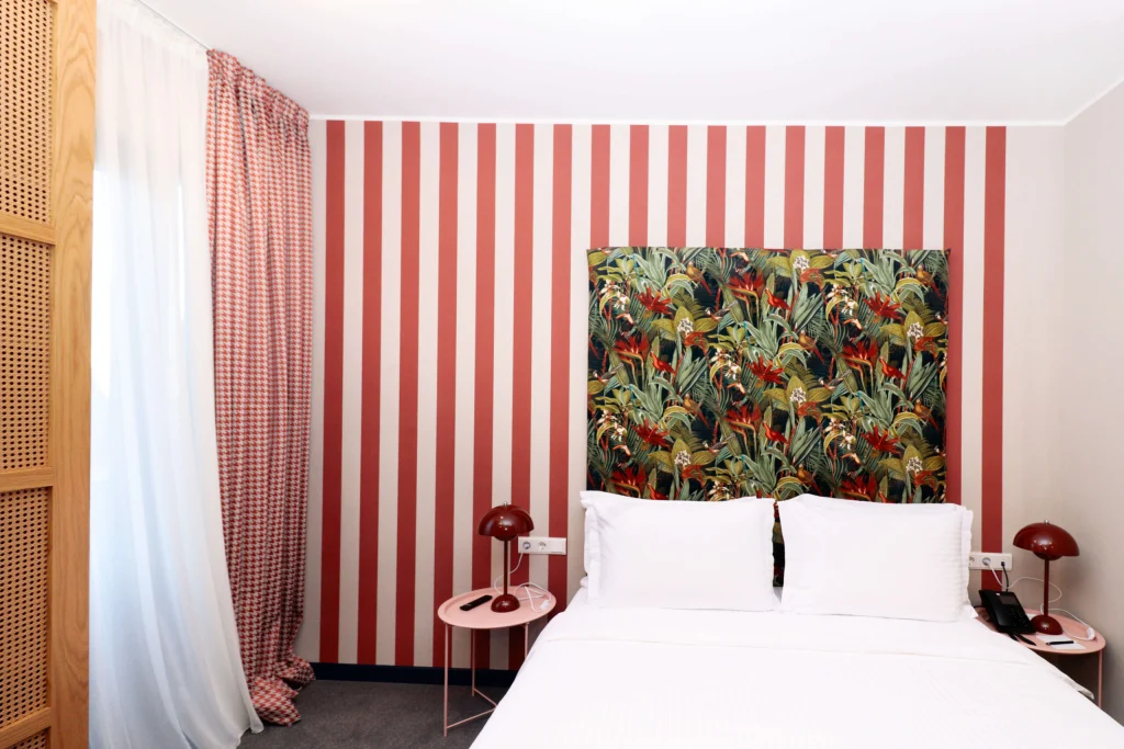Margot Sololaki — Bedroom with bold red striped wall, tropical-print headboard, and white bedding.