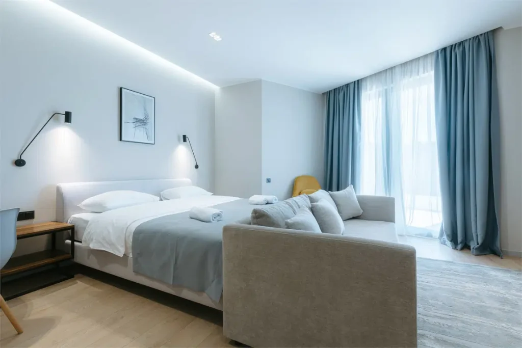 Mziuri Gardens — Bright bedroom with a bed, sofa, and large window framed by blue curtains.