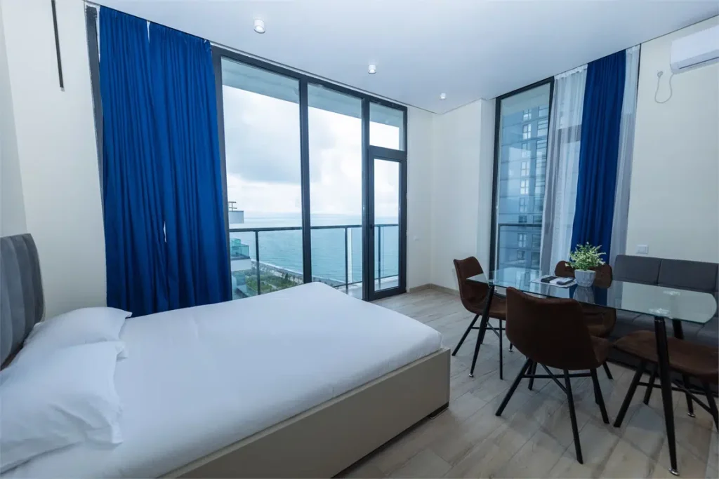 NEW WAVE APART-HOTEL — Guest room with sea-view balcony, blue curtains, bed, and small dining table.
