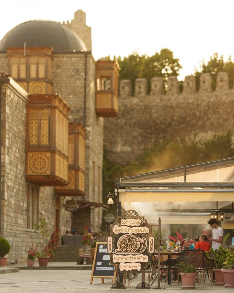 Natenadze Wine Restaurant — Exterior view of the stone building with ornamented balconies and ancient fortress wall behind.