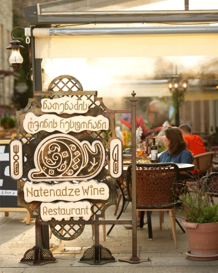 Natenadze Wine Restaurant — Outdoor restaurant terrace seating with awning, guests dining under warm evening light.