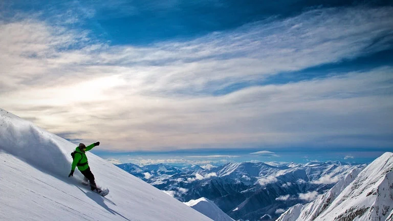 New Gudauri Rooms — Snowboarder descending a slope with panoramic mountain views in the background.