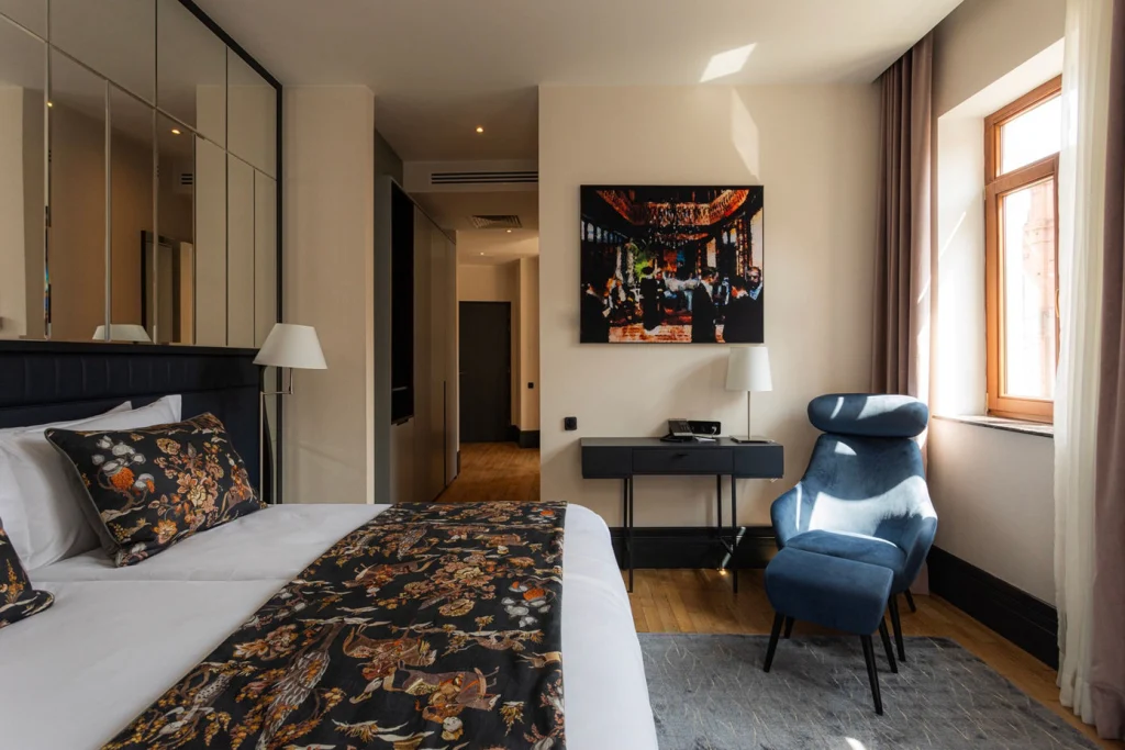 Puris Moedani Boutique Hotel — Stylish bedroom with patterned bedding, blue armchair, and large window.