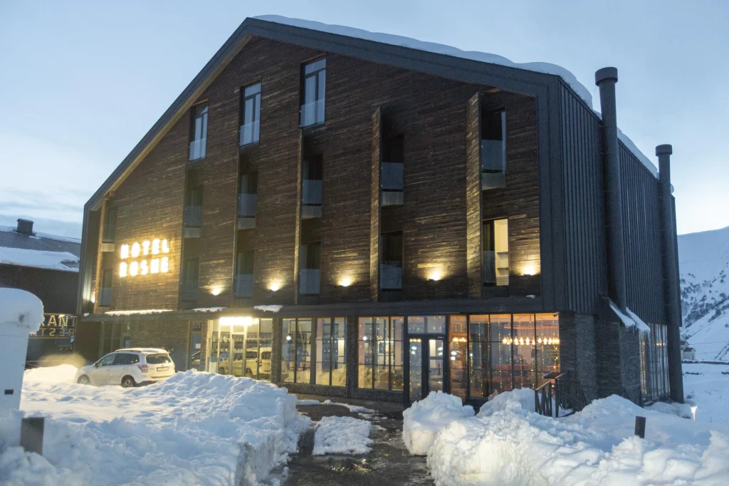 Roshe Gudauri — Wooden-clad hotel exterior illuminated at dusk with snow banked along the entrance.