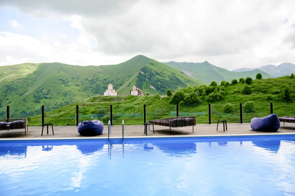 Roshe Gudauri — Outdoor pool deck with loungers overlooking green mountains and a small church.