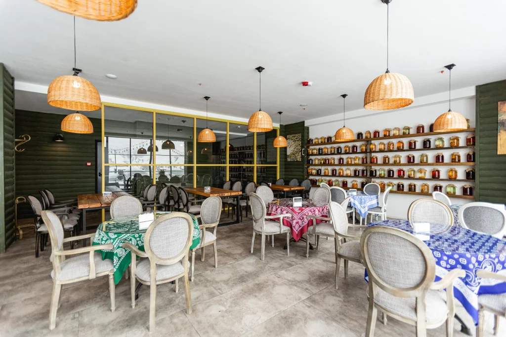 Roshe Gudauri — Hotel restaurant dining room with patterned tablecloths and shelves of preserved jars.