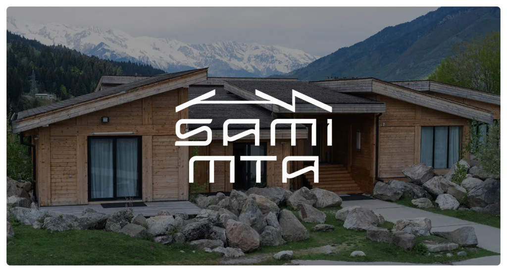 SamiMta — Wooden lodge-style building with mountain views, surrounded by large rocks and greenery.