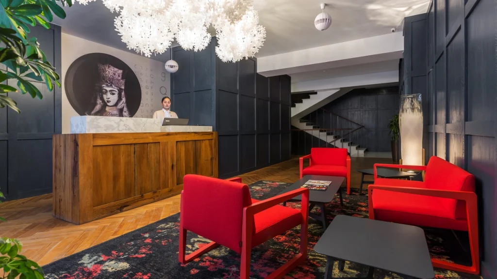 Shota@Rustaveli boutique hotel — Hotel lobby with wooden reception desk, red armchairs, and decorative pendant lighting.