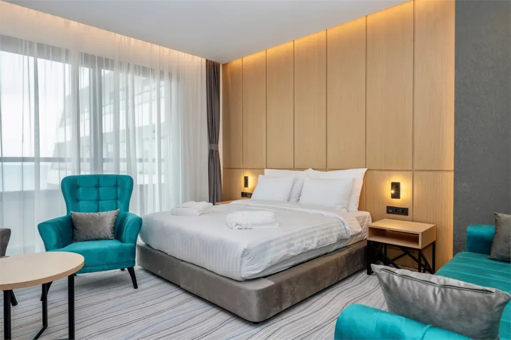 The Grandeur Hotel — Modern hotel room with a large bed, teal armchairs, and natural light from windows.