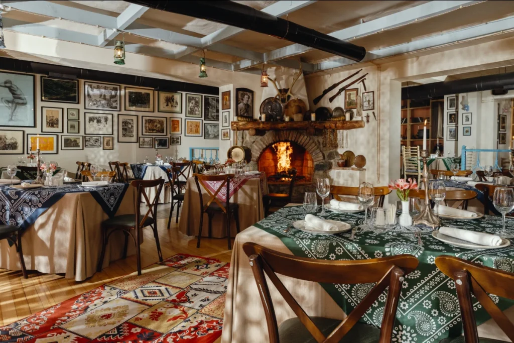 Tsiskvili in Tskneti — Rustic dining room with fireplace, framed pictures, patterned rugs, and set tables.