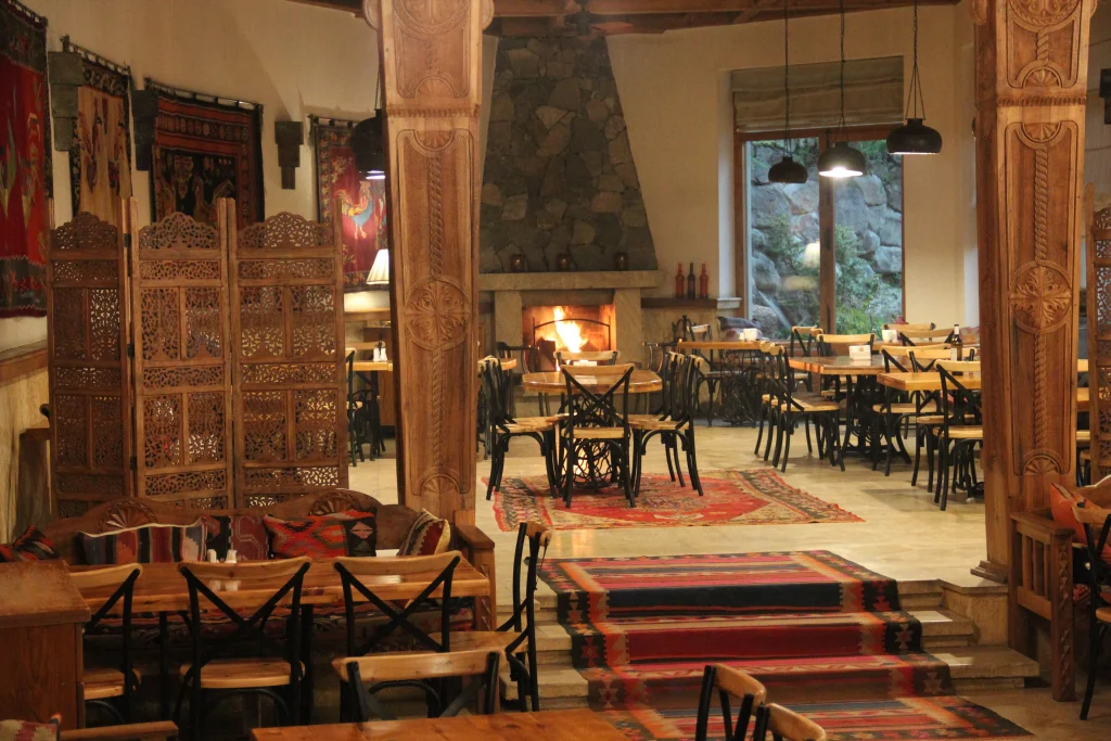Vardzia Resort — Rustic restaurant interior with carved wooden pillars, rugs, and a stone fireplace.
