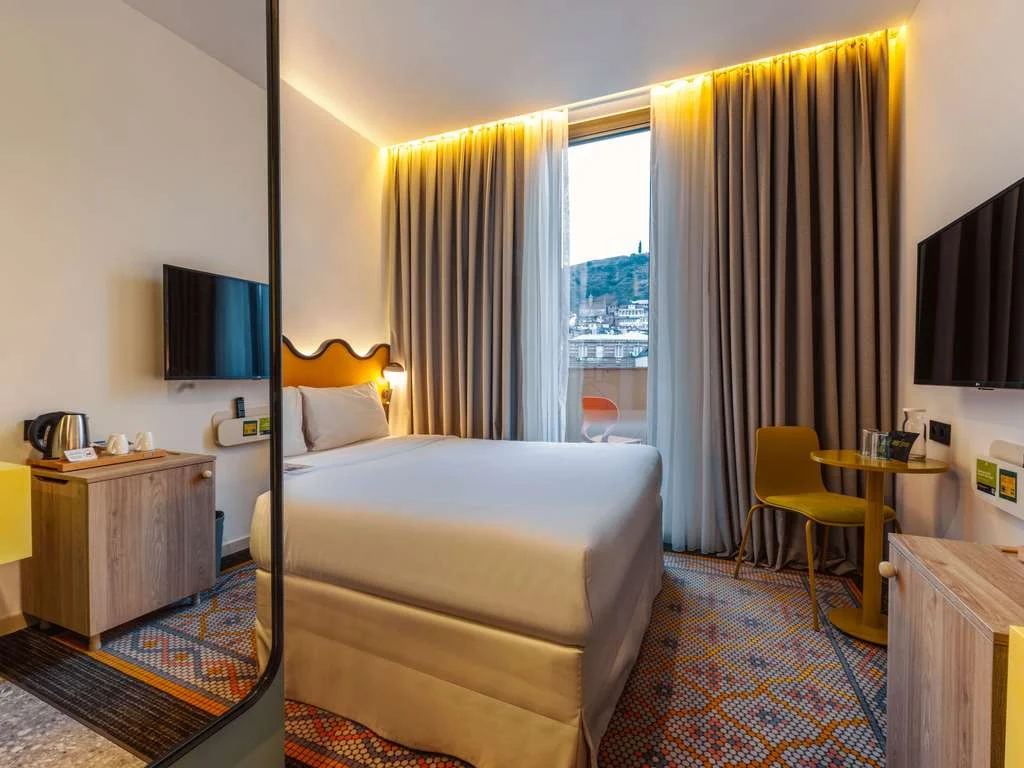 ibis Styles Old Tbilisi — Hotel room with a bed, seating area, and window overlooking the city.