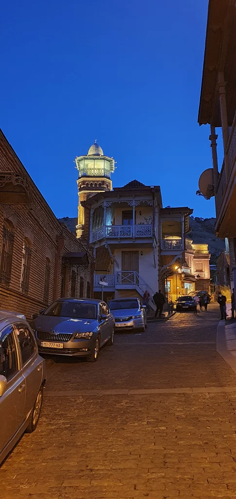 An evening street in Old Tbilisi with traditional balconies and a lit minaret.